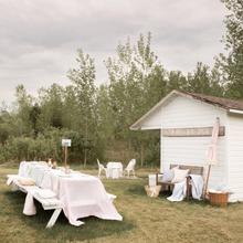 Load image into Gallery viewer, Outdoor setting with white tables and chairs in a grassy area near a small building.