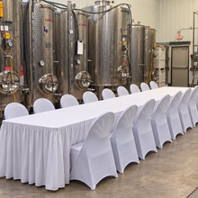 Load image into Gallery viewer, A long banquet table covered in a white tablecloth with matching white chair covers, set in a brewery or industrial kitchen with large stainless steel fermentation tanks in the background.