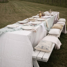 Load image into Gallery viewer, Dining table set for a meal outdoors with plates, cutlery, and decorative pillows on a grassy field.
