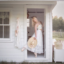 Load image into Gallery viewer, Woman in a white dress holding a straw hat, standing in a doorway of a white building.
