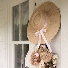 Load image into Gallery viewer, Woven straw hat with a pink ribbon hanging on a white door with floral decorations.
